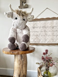 Plush highland cow toy on a wooden table in a cozy bedroom. The highland cow has tan fur and crochet horns, ears, and feet.