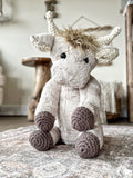 Plush highland cow sitting on a carpet in a nursery. The highland cow stuffed animal has soft tan fur with crochet horns, ears, and feet.