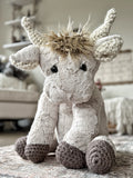 Plush highland cow with soft tan fur is sitting on a carpet. The highland cow has crochet horns, ears, and feet.