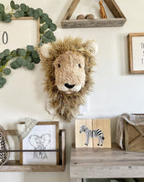 A plush lion head made by ClaraLoo that is mounted on a white wooden plaque in a safari themed nursery. The stuffed lion has soft tan fur, a shaggy brown mane, and crocheted accents.